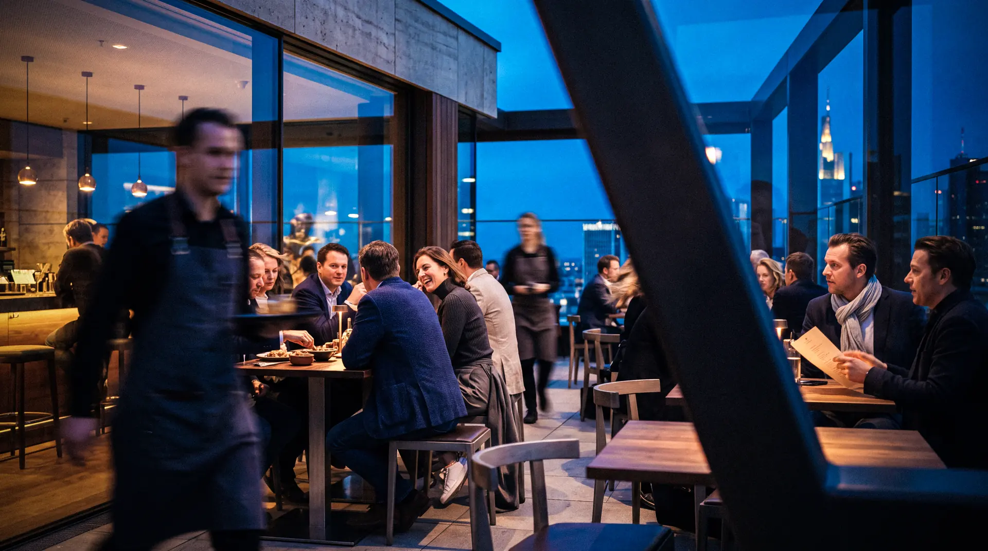 Eine Dachterrasse in der blauen Stunde, Gäste an Tischen vor der Skyline.